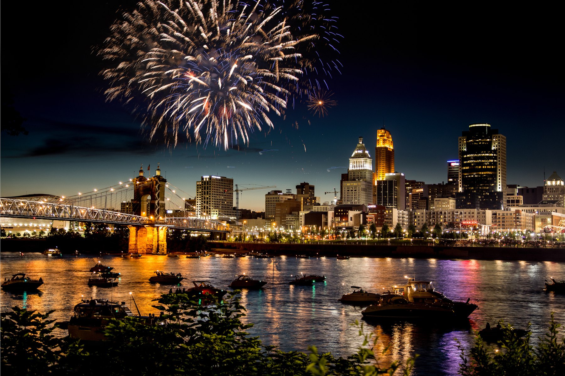 Fireworks over the Ohio River and the downtown Cincinnati skyline, with the John A. Roebling Suspension Bridge lit up and boats gathered on the water — September 2026 events across Butler, Warren and Hamilton counties, Kristine Green Realtor