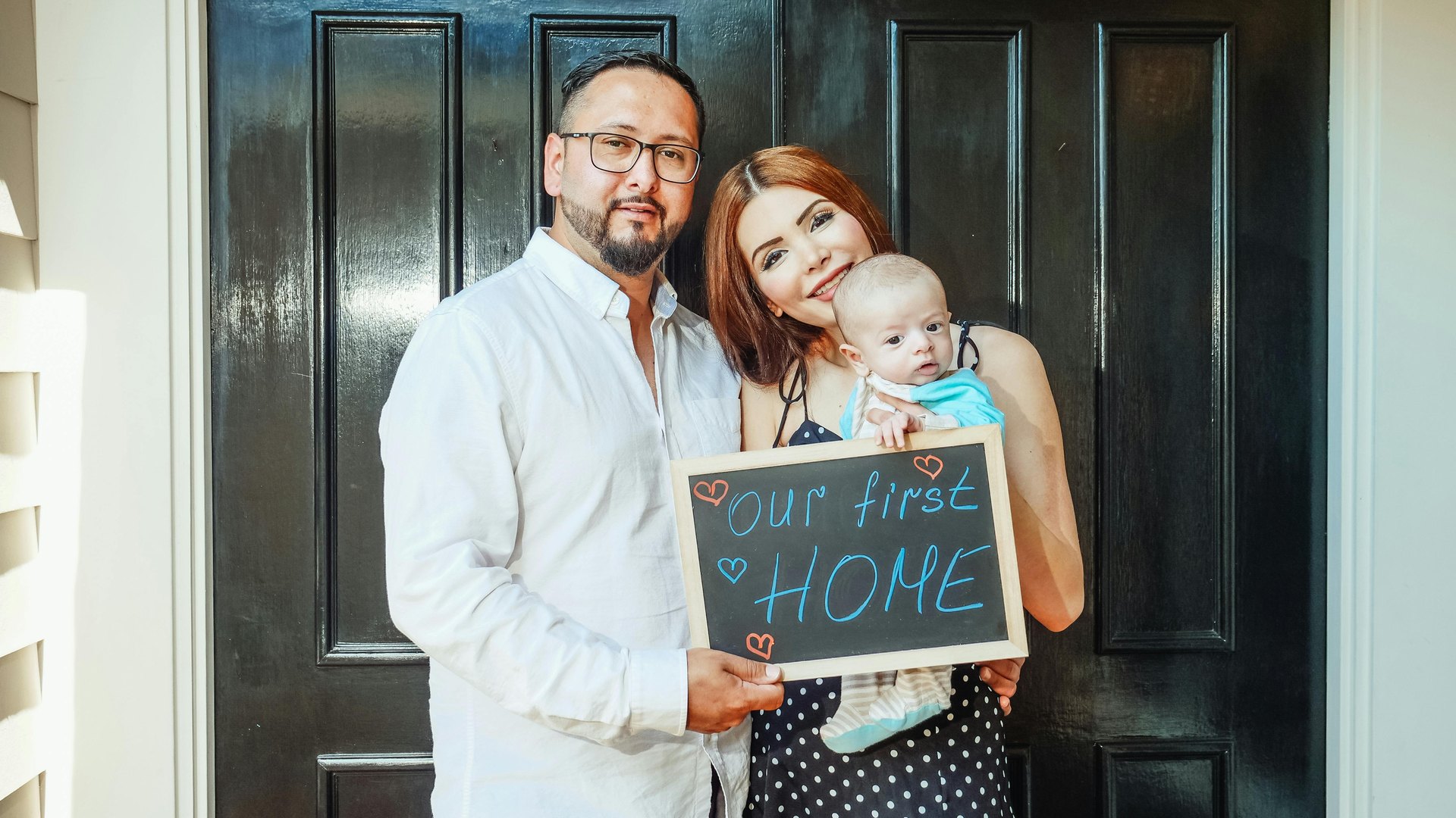 Couple holding a baby and an Our First Home chalkboard sign at their front door after saving for a down payment in Ohio, Kristine Green Realtor